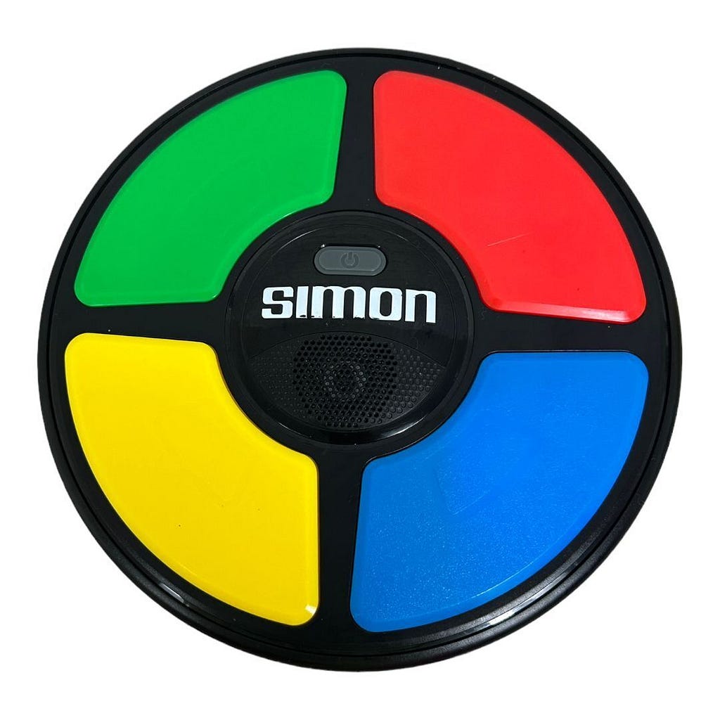 The classic Simon electronic memory game was a disc-shaped toy about the size of a medium pizza and a few inches tall. There is a black circle in the center that reads “Simon.” The remaining surface area is divided into four sections creating a sort of wheel around a hub appearance. Each section is a different color. Starting at the 12 o’clock position and moving clock-wise, the colors are red, blue, yellow, and green.