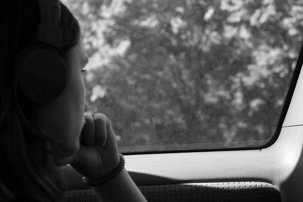 Woman, introspective, wearing headphones, looking out car window with a lonely expression on her face, hand on chin.