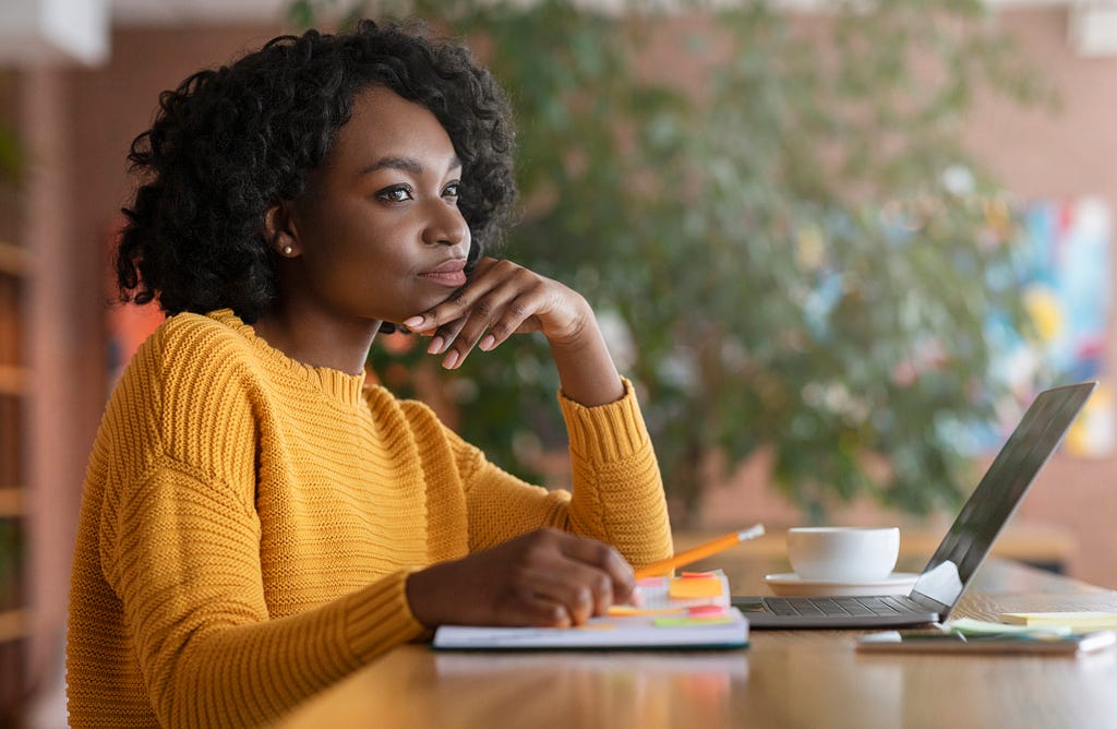 A woman sits with her hand on her chin and a pencil in her hand. She has a notebook in front of her.