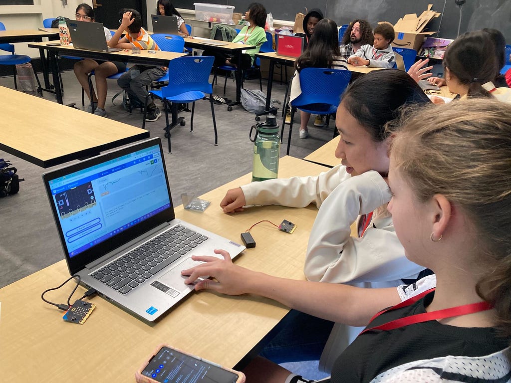 Photo of a middle school classroom with a dozen students coding on laptops at different tables. A mentor is speaking with 3 students in the back.