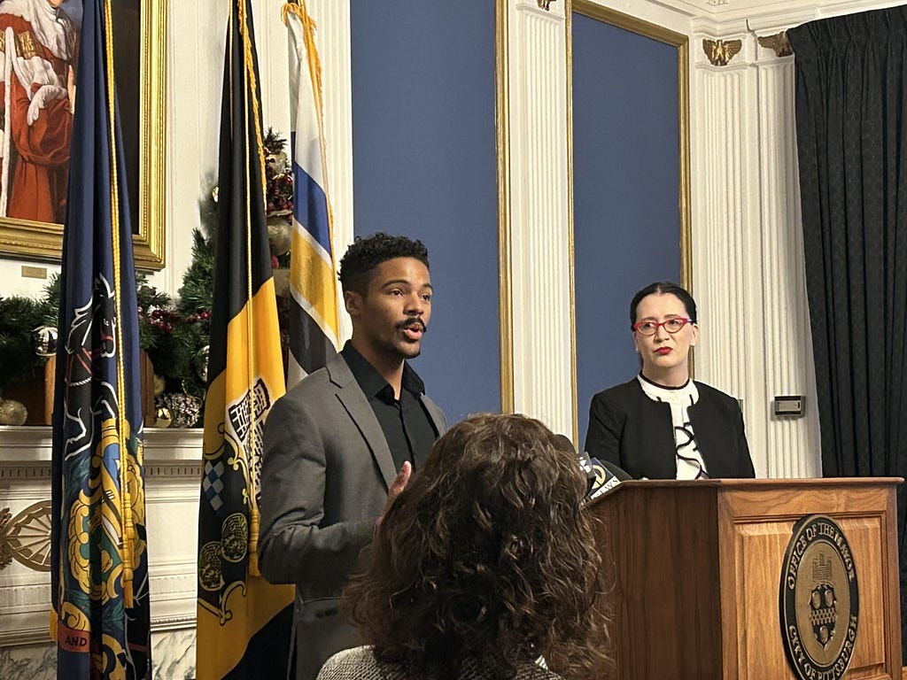 A person standing and speaking at a podium in an ornate room while another person stands, listens, and looks on from the right side.