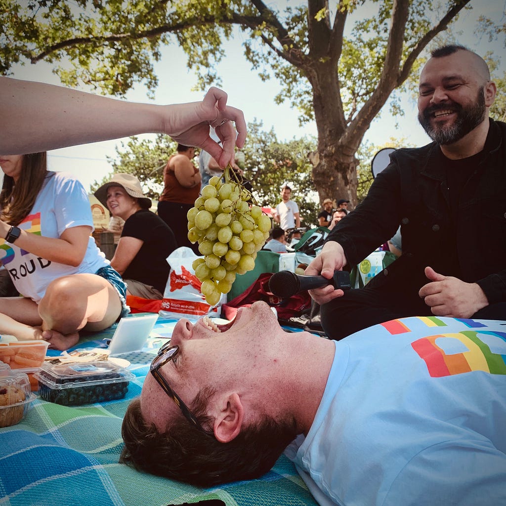 The author laying down on a picnic blanket, while being hand-fed grapes.