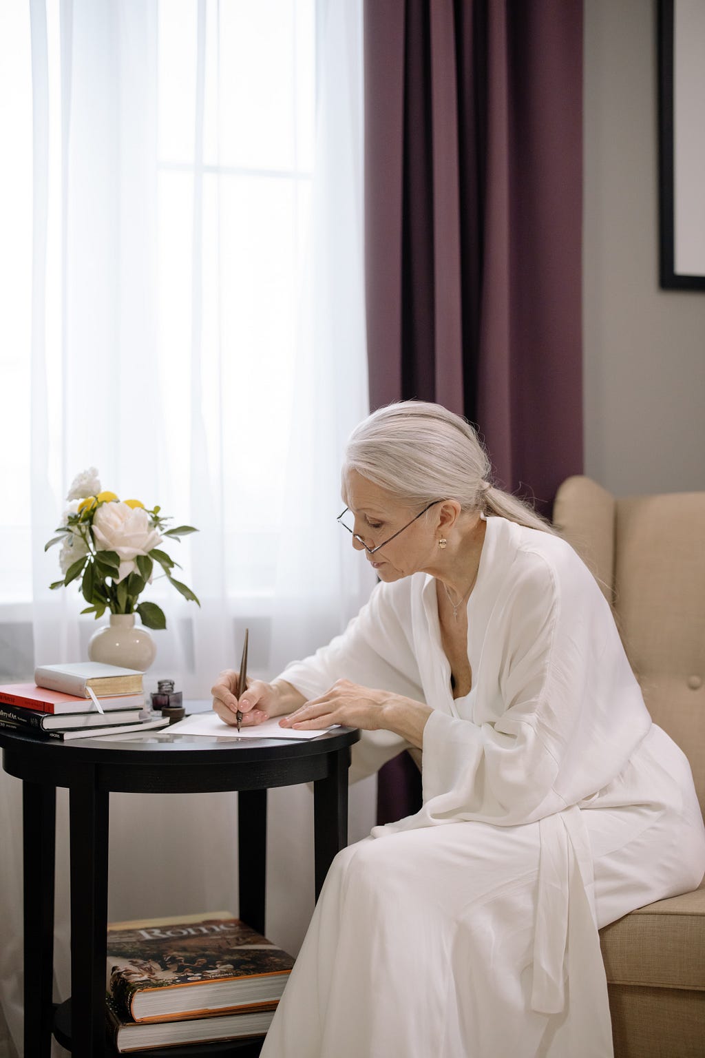 Grey haired woman in a white robe, sitting at a desk, intently writing on a piece of paper with a fountain pen.