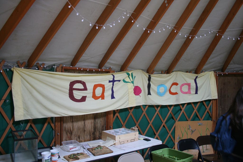 A cloth, hand painted sign reading “eat local” displayed over a table of food