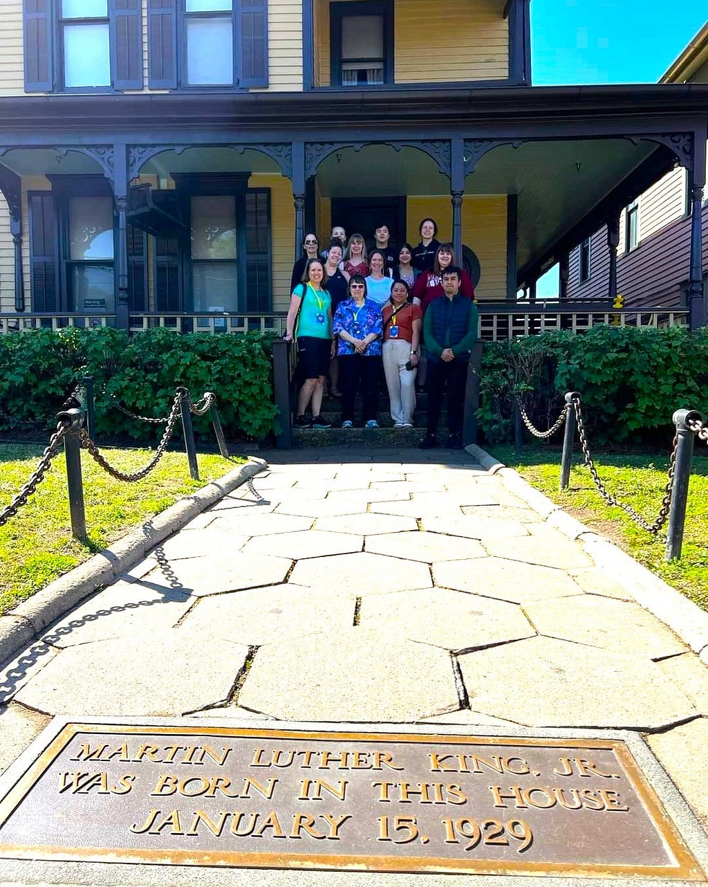 A group of 13 people are posing on the steps of an old Victorian-style home. On the stone pathway in front of them is a plaque saying Martin Luther King Jr. lived in the home.