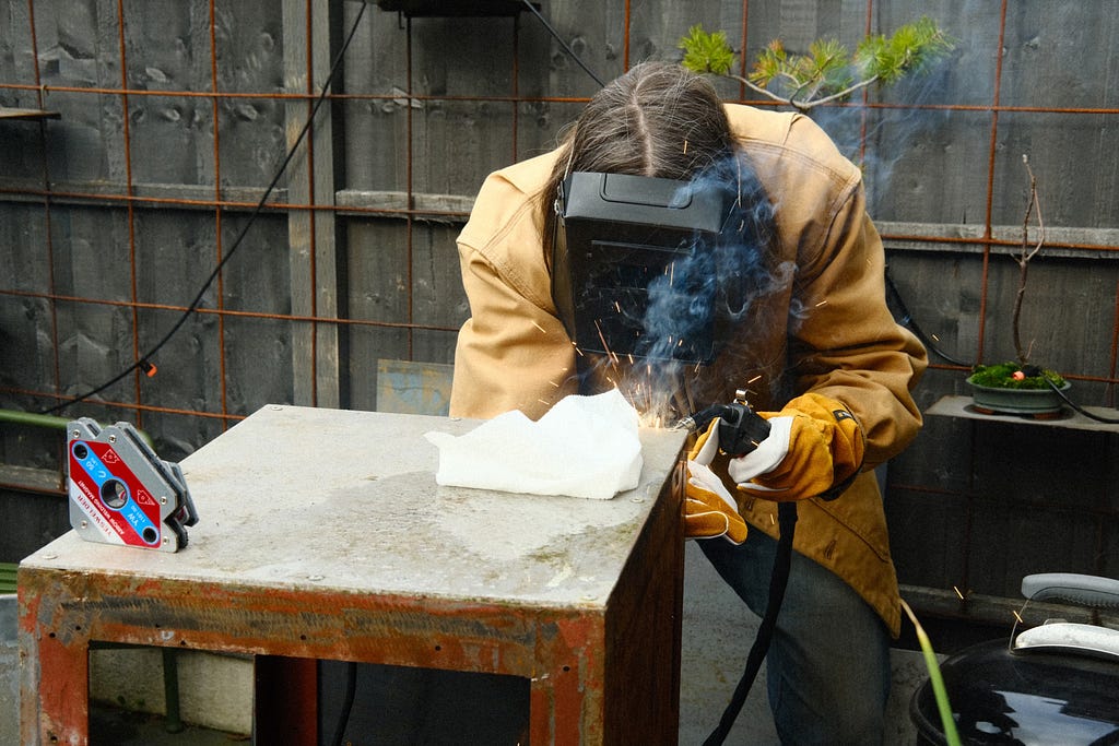Rebecca with a welding torch and mask on, welding a metal box
