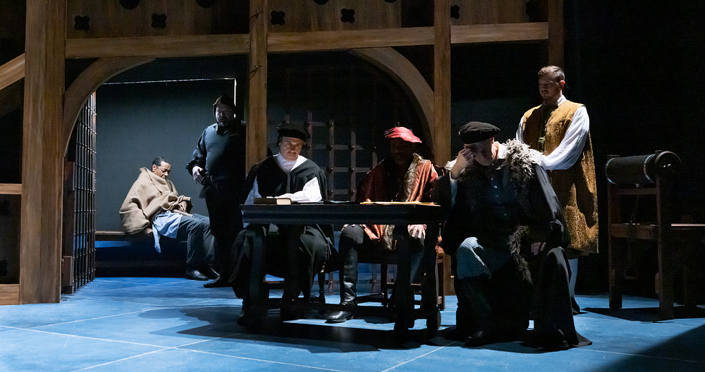 A man sits in a prison cell under a brown blanket guarded by a jailer, while four men in 16th century clothes sit at a table outside, looking frustrated.