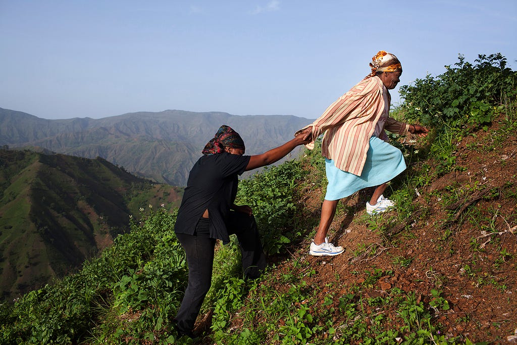 Two women climb up a hill with reaching back to help the other ascend a steep step.