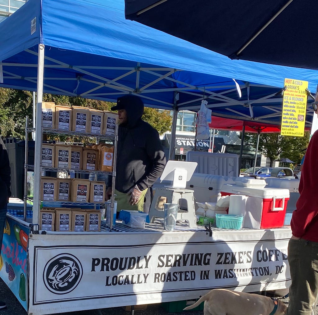 Image of a Zeke’s Coffee farmers market stall with packages of coffee for sale on the left, register center, and a banner with the words, “proudly serving Zeke’s Coffee” below.