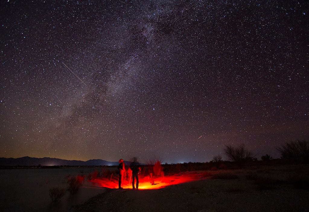 Two people stand in a circle of red light, looking up at the night sky with millions of stars and the Milky Way.