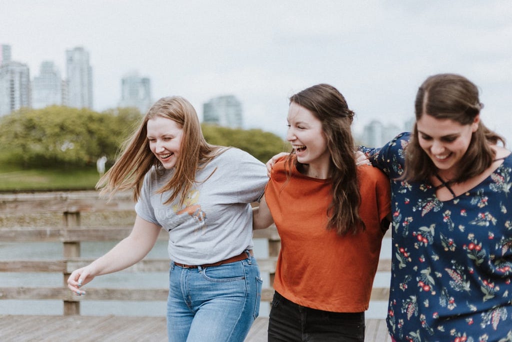 A photo of three young women walking along while laughting and having their arms around each other.