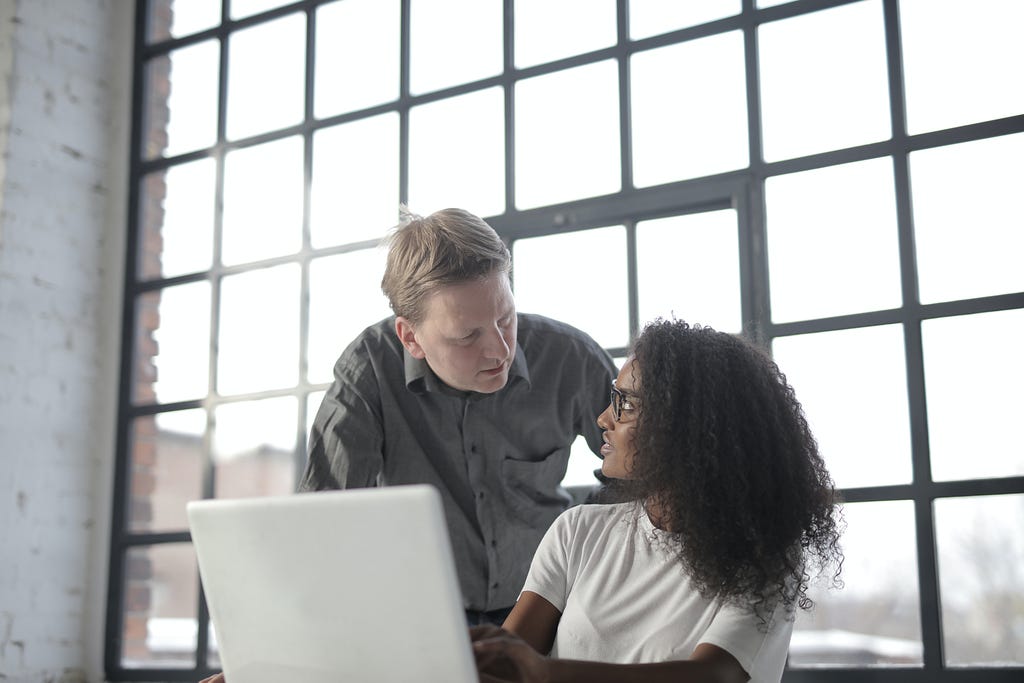 Two people having conversation in front of a laptop. They appear to be in a room with huge window glass window on the background. Photo by Andrea Piacquadio: https://www.pexels.com/photo/focused-multiracial-coworkers-of-different-age-discussing-business-issues-3865843/