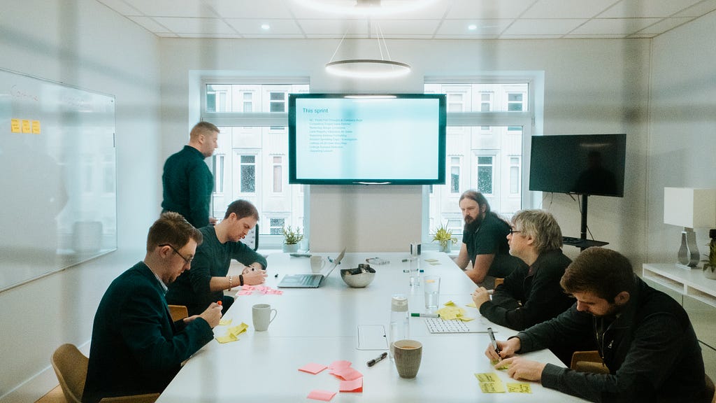 A team of highly-focused developers, sit around a long white table, covered in multi-coloured post-it notes, planning a new development sprint.