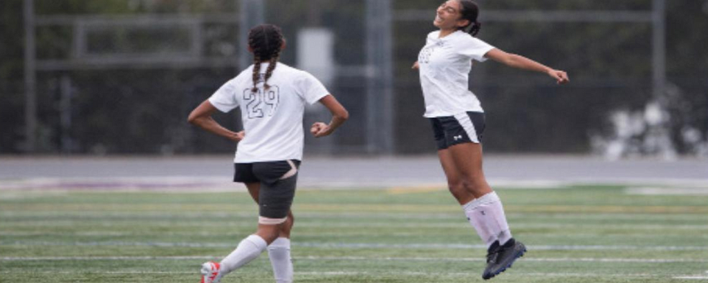 Photo of the Traylor sisters on a green soccer pitch. They are wearing white jerseys with black shorts and white shin guards. One of the sisters is jumping in the air.