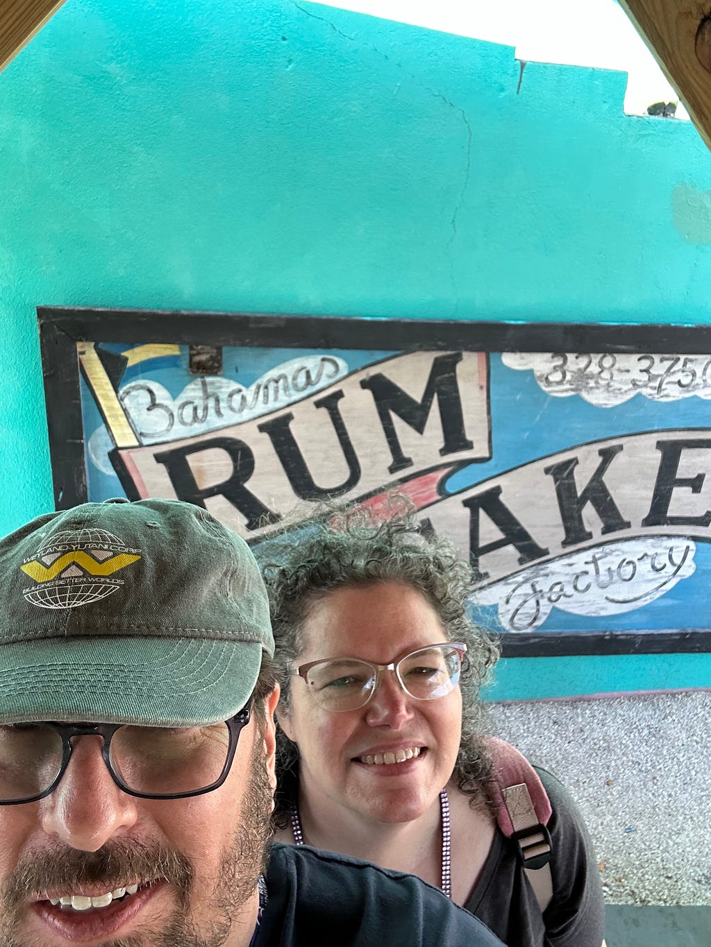 A middle aged couple stands in front of a painted sign advertising the Bahamas Rum Cake Factory. The painted sign has a blue background with clouds behind the words “Bahamas”, “Factory” in cursive font, and the store’s phone number, a small Bahamian flag, and the words “Rum” and “Cake” banner style in capital letters.