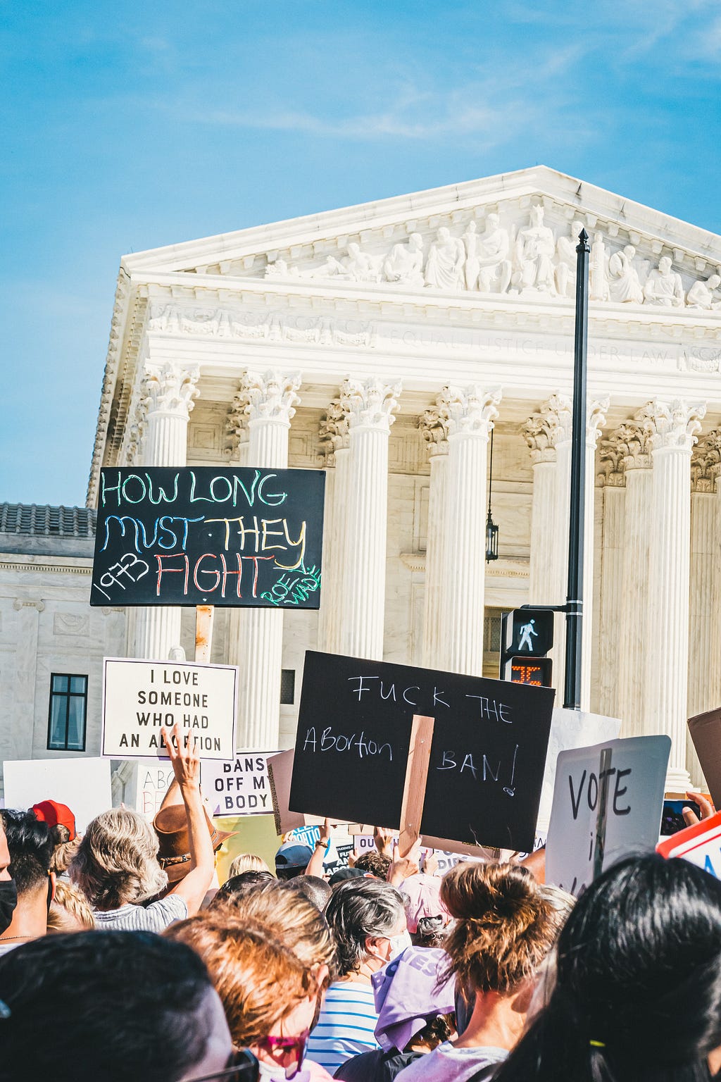 Image of protestors in front of the Supreme Court with signs reading “How long must they fight. Roe Wade,” “Fuck the abortion ban,” and “I love someone who had an abortion.”