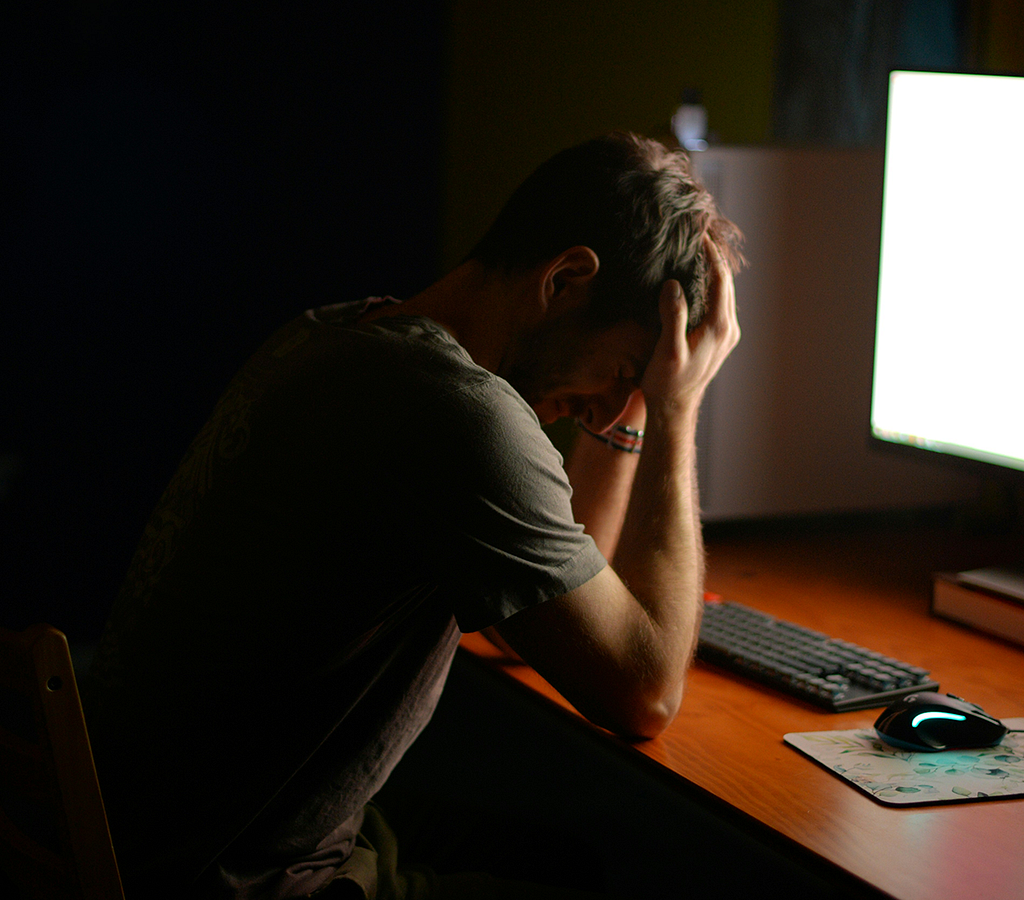 A distressed man in a dark room in front of a computer with their head in their hands.