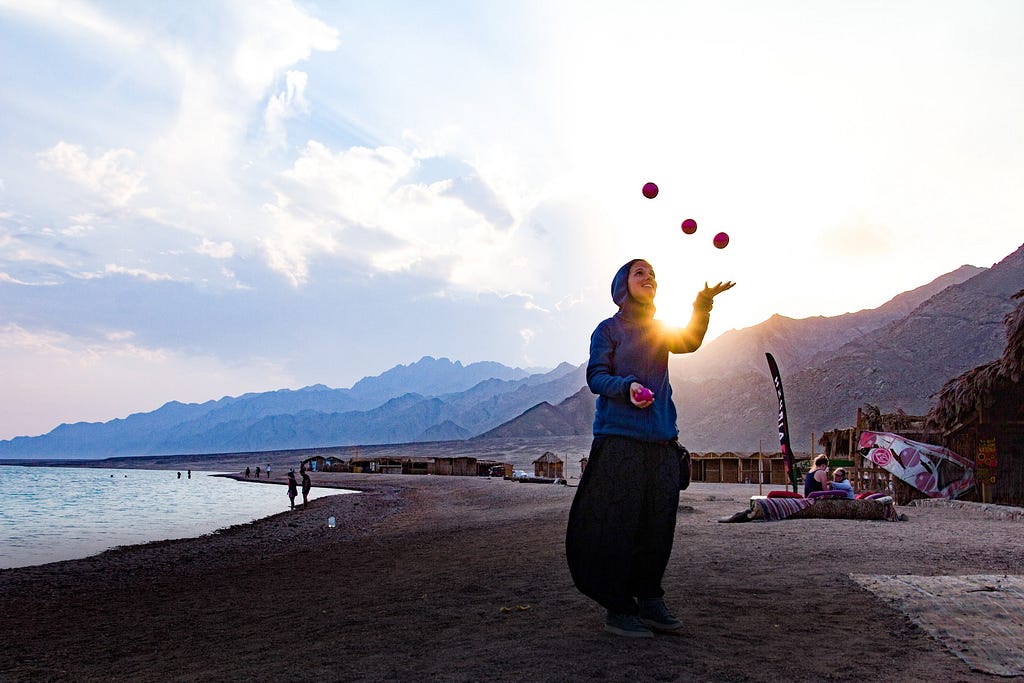 Woman standing by a lake smiling and juggling balls.