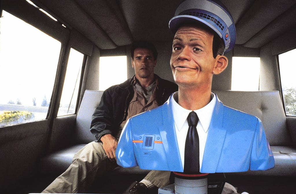 A torso of a plastic-looking male figure in a light blue suit and hat and a black tie, driving a taxi with Arnold Schwarzenegger sitting on the back seat behind the robotic taxi driver.
