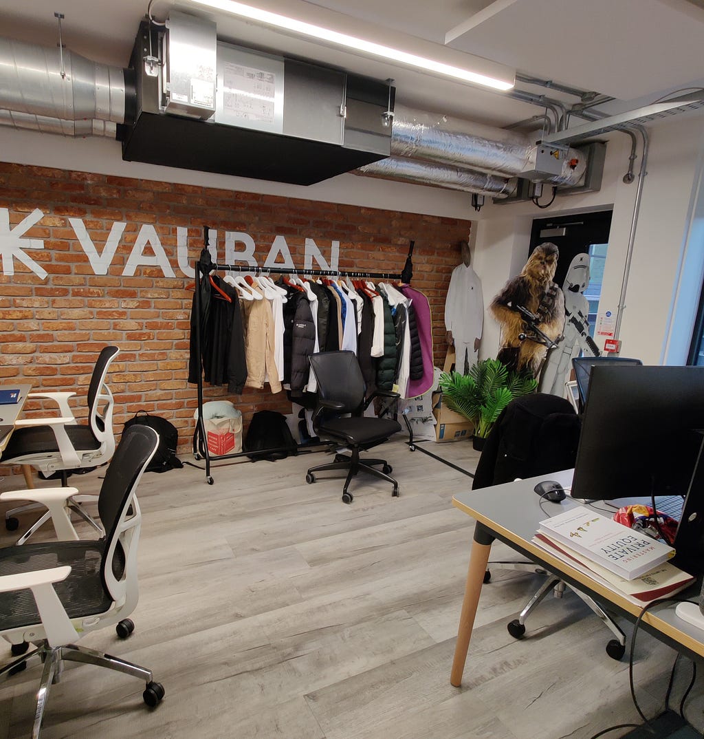 a corner of the office with desks, coats, and the vauban logo on a brick wall