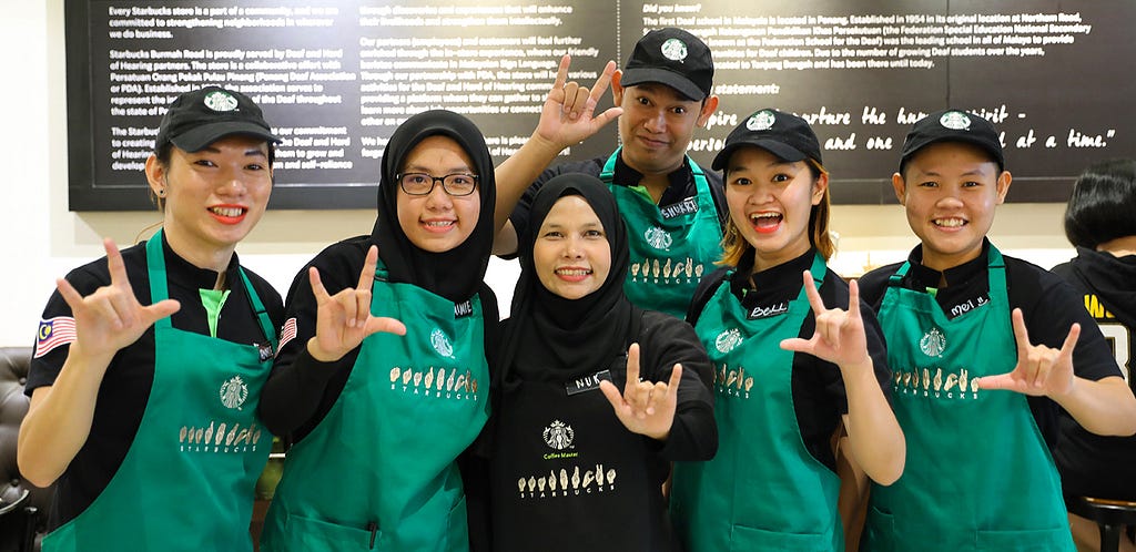 A group of Starbucks staffs from the Starbucks Signing Store in Penang posing for a group photo. The team are wearing the Starbucks Signing apron and hand gesturing ”rock on”.