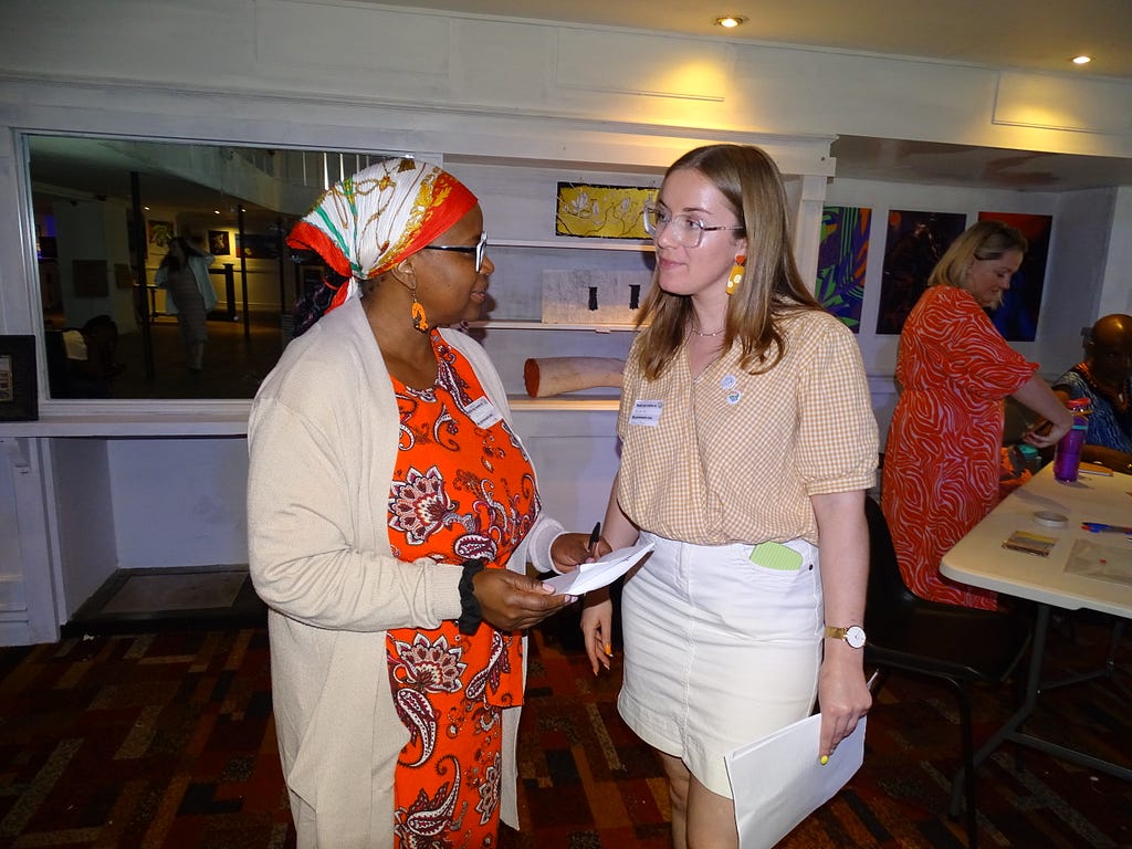 A black woman with a red dress and headscarf stands on the left, talking with a white blonde woman with a yellow top and white skirt on the right.