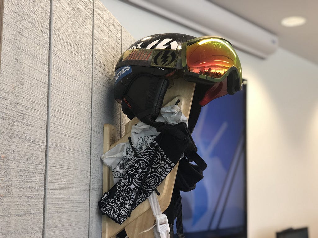 The Strax Rack hanging on a false gray wall with a snow helmet, goggles, and bandanas hanging it from it to show how it works