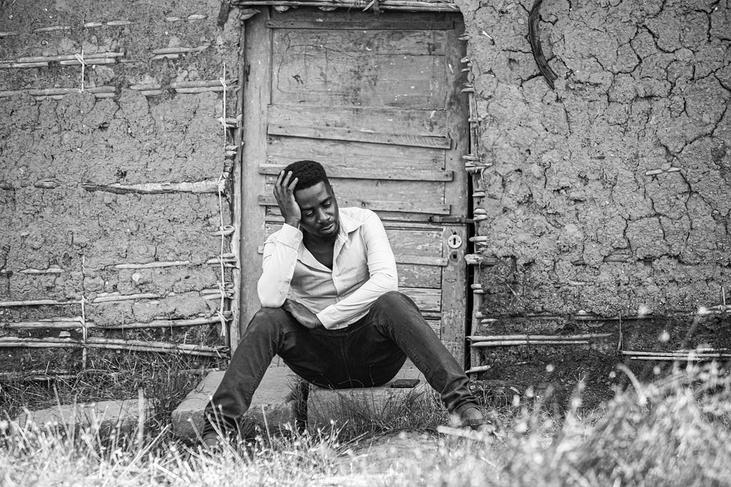 A black man in front of an old shack sitting at door with one hand on his head