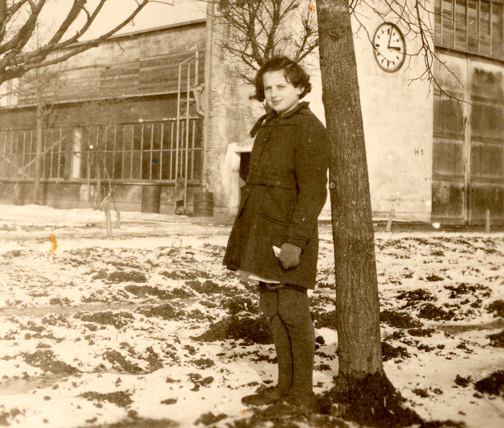 A girl stands next to a tree. She is wearing a heavy coat, mittens, and stockings. There is snow on the ground. In the background, there is an industrial building with a large clock in the wall facing the camera.