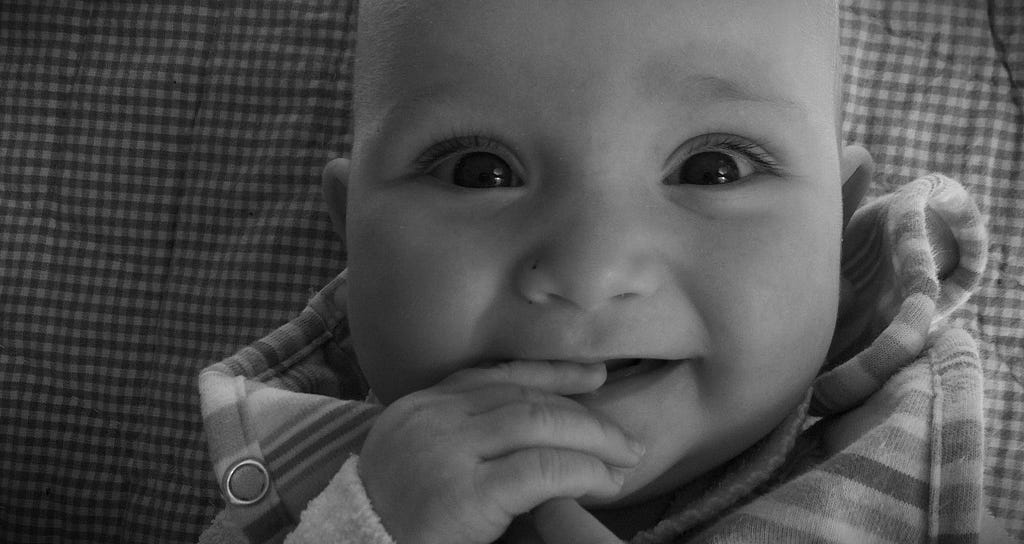 a young white skinned baby smiles directly into the camera. The baby’s eyes are sparkling with laughter. It is a close up headshot. You see the baby has their right hand in their mouth, and they are smiling but don’t yet have any teeth. They are wearing something stripy, and lying on a plaid blanket.