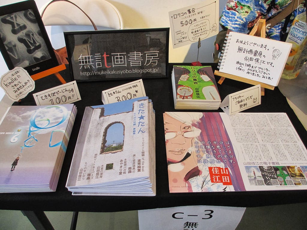 A booth at a book event in Fukuoka, displaying various books, pamphlets, and a Kindle device. The booth features a sign that reads ‘無計画書房’ along with information about the books and pricing. Several books are displayed prominently on the table.