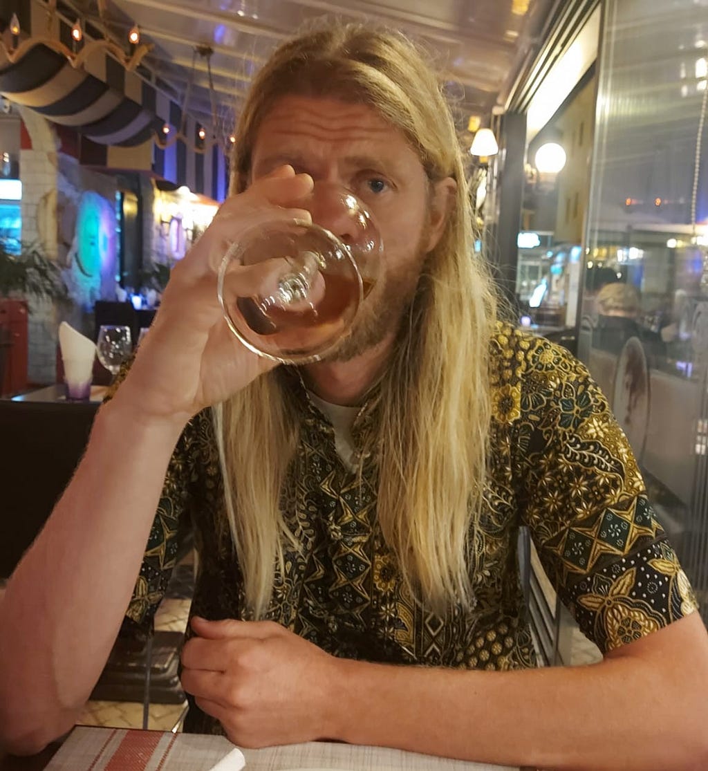 Man with long, blonde hair, wearing a green and yellow patterned short-sleeved shirt drinking from a glass hiding most of his face. 
 .