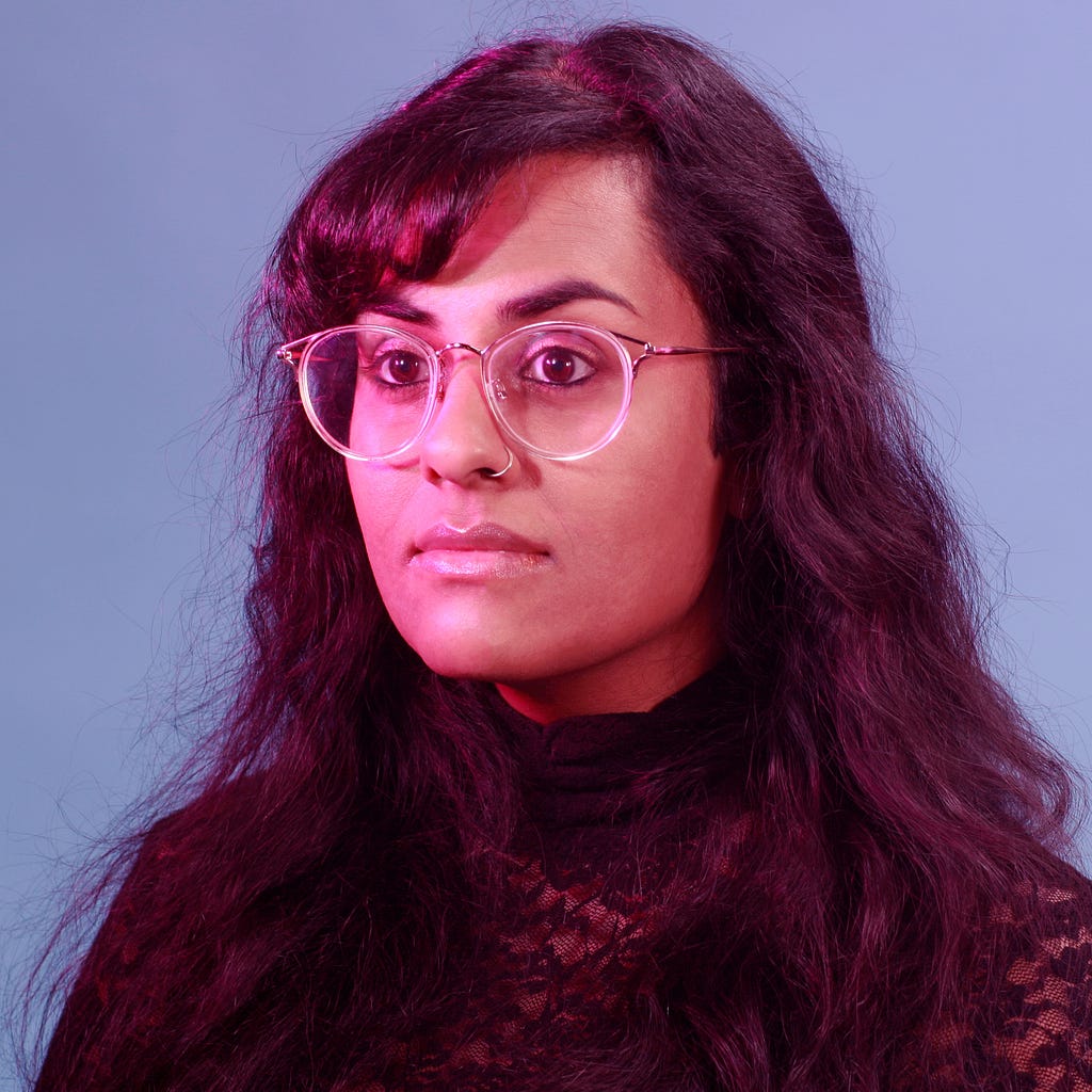 A brown-skinned woman with long black hair, wearing a black turtleneck and clear glasses. She sits against a blue backdrop.