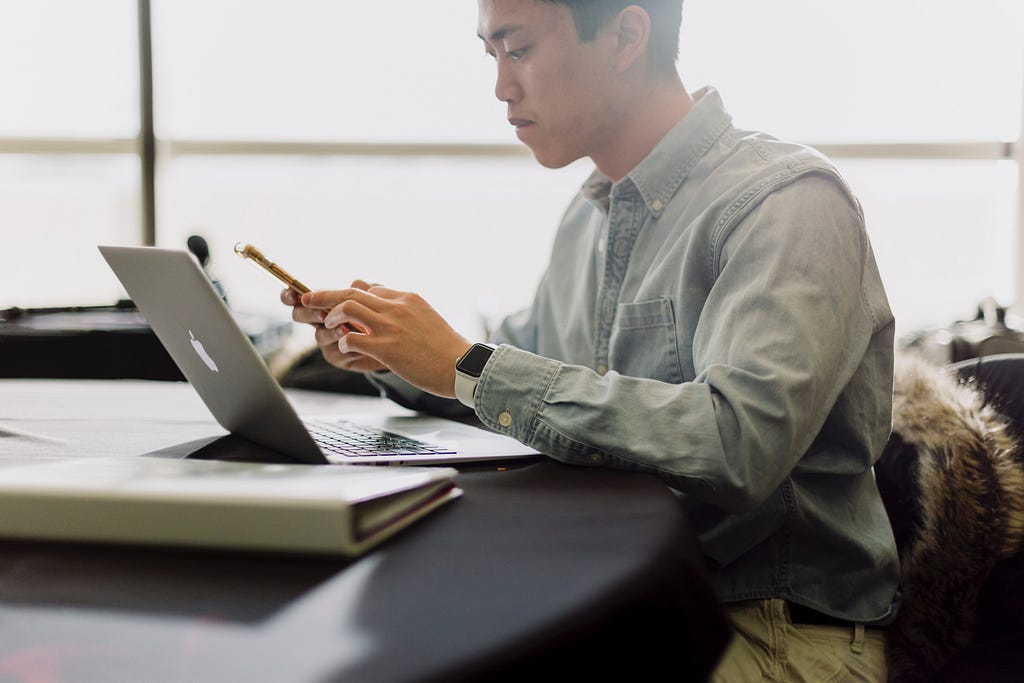 A man wearing a button-down shirt and sitting at a desk begins to make a call on his cell phone.