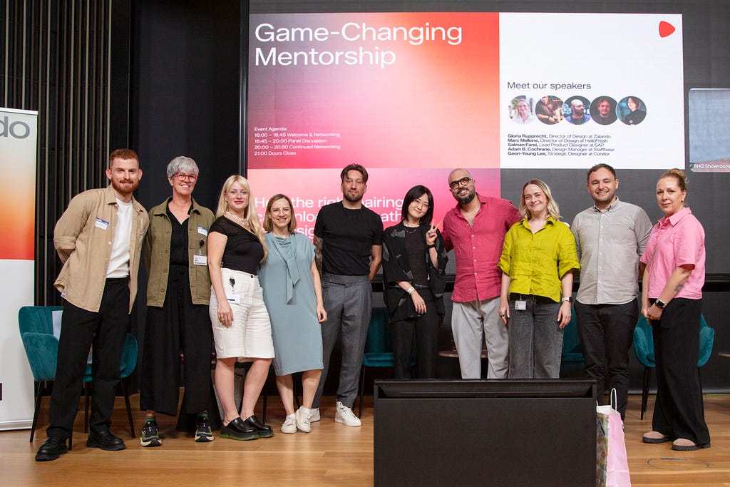 A group of 10 people standing in a line, facing the camera and smiling, with a large screen behind them that reads “Game-Changing Mentorship”.