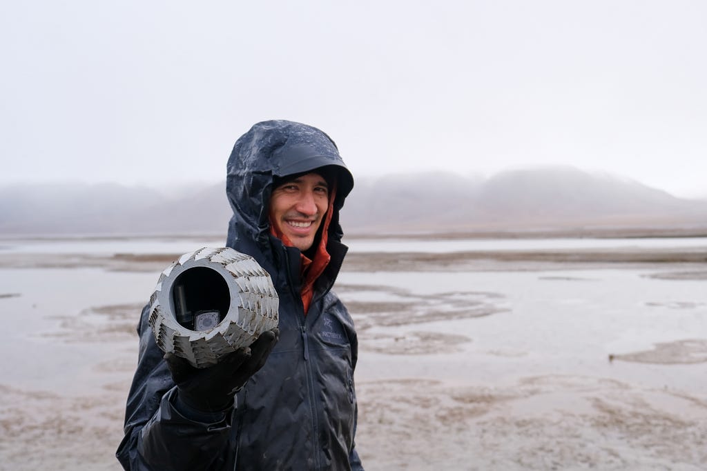 Joe Kennedy wearing a black winter jacket with the hood over his head, holding a grey hollow orb towards the camera. Misty brown and grey landscape in the backdrop.