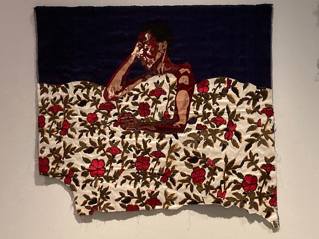 A photograph of an embroidered picture of a Black woman sleeping beneath a white quilt decorated with red flowers and green leaves.