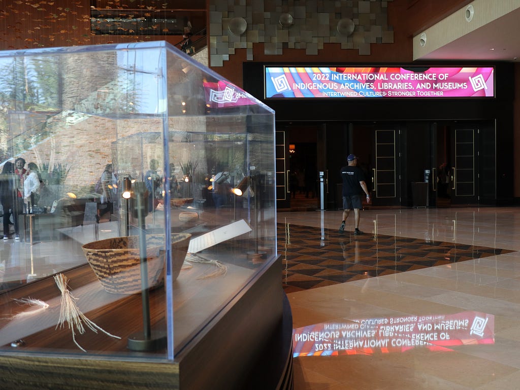 Photograph shows a large hotel lobby with shiny floor, and long illuminated sign showing entrance to a conference venue. In the foreground is a glass museum case showing a woven basket.