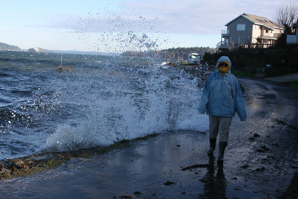 A street on the waterfront with a wave crashing up against it. A woman in heavy coat and boots walks toward the camera. There is a house visible behind her.