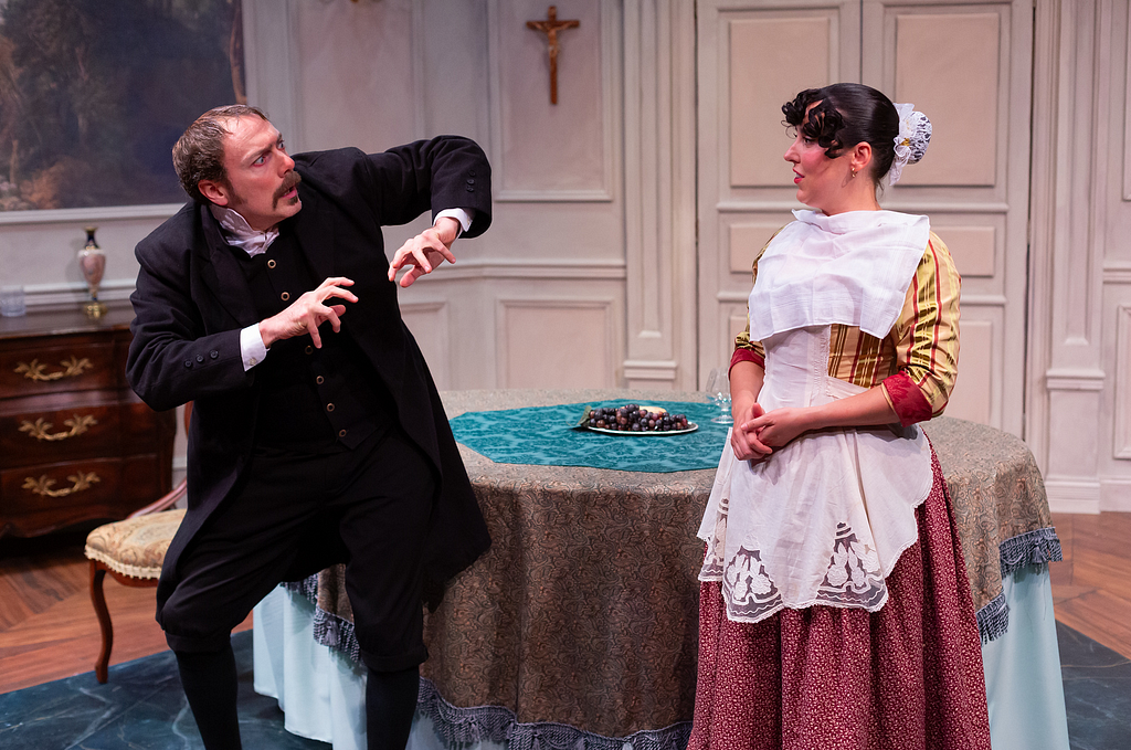 A man in black 19th century clothes recoils at a smiling maid with a napkin over her chest.