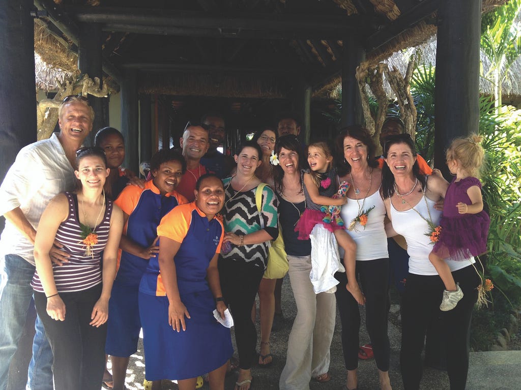 Kelley Steckelberg and a large group of adults and children smile under an overhang, with lush greenery nearby