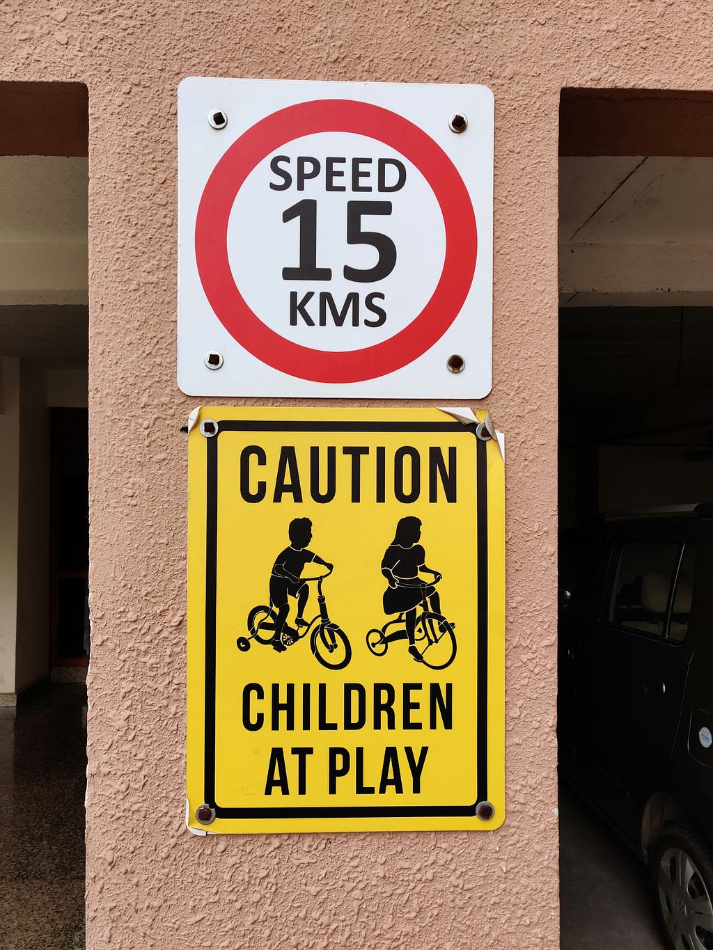 Two signs mounted on a textured peach-colored wall: a circular red and white speed limit sign reading ‘SPEED 15 KMS’ above a rectangular yellow caution sign with black text and silhouettes of two children riding bicycles with training wheels, reading ‘CAUTION CHILDREN AT PLAY