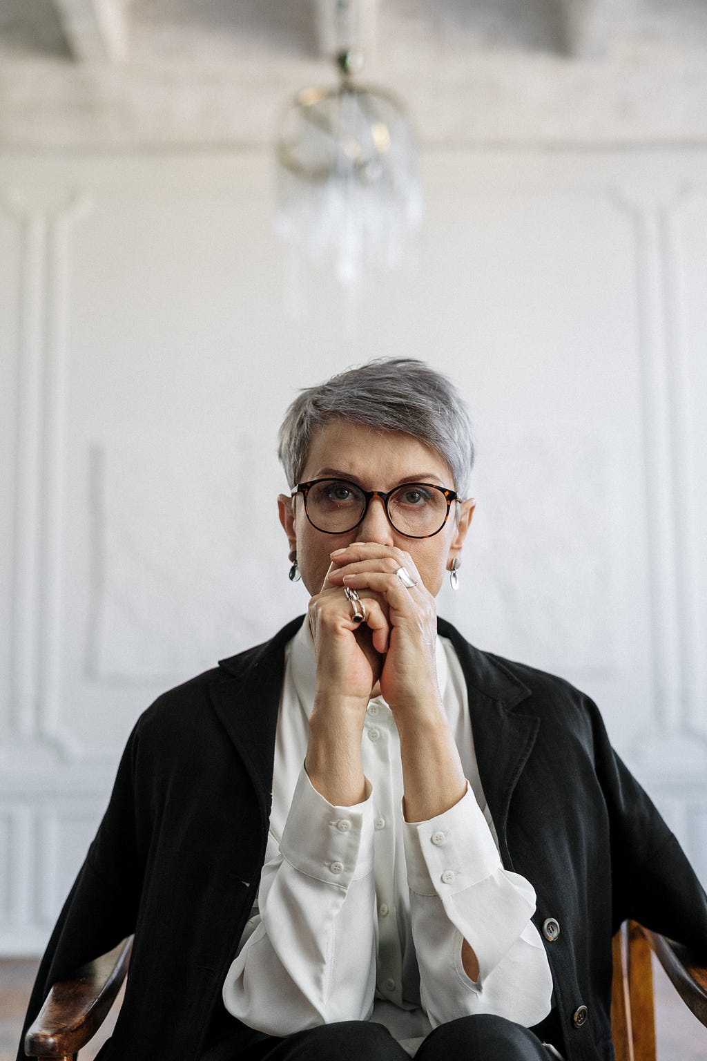 A woman in her late 50s/early 60s stairs straight at the camera looking stern with her hands folded in front of her mouth.