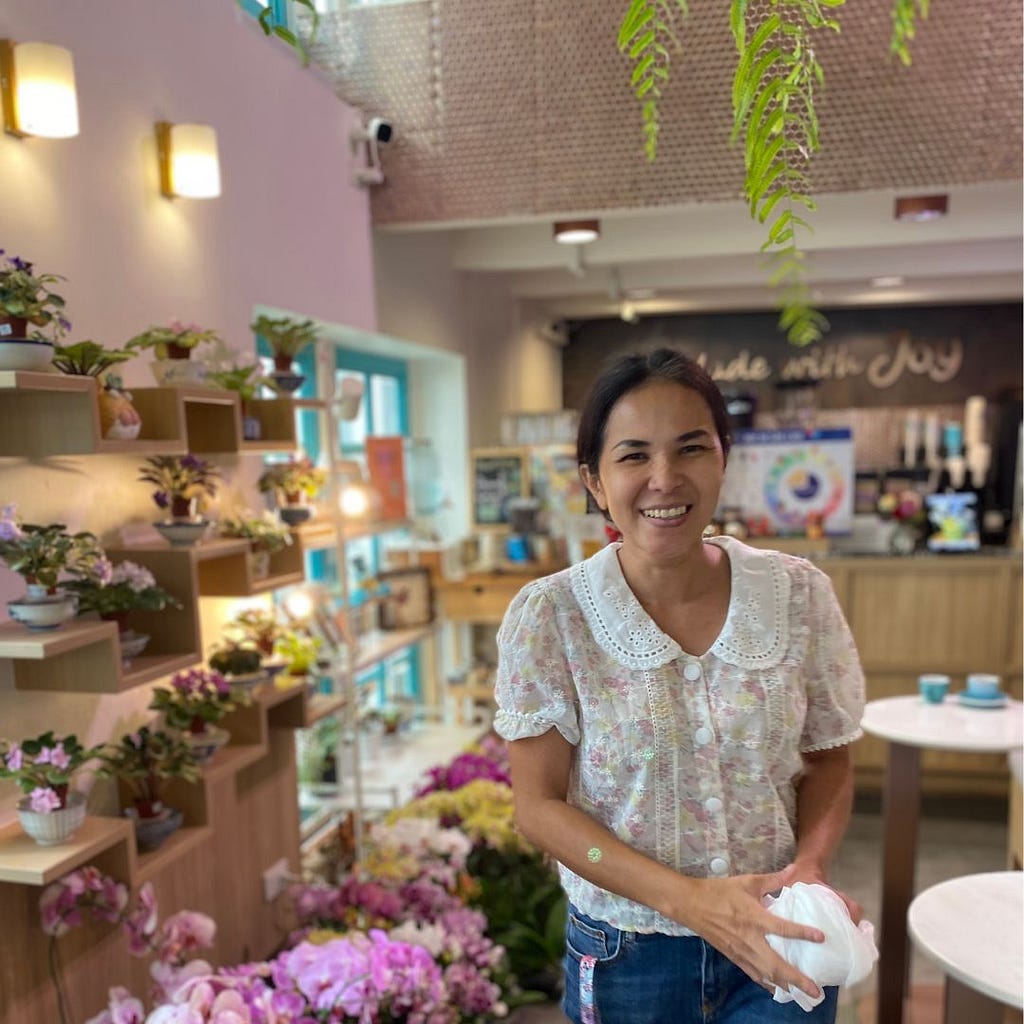 Thai woman in white blouse walks towards camera smiling, plants line shelves on left and blackboard in background reads ‘made with joy’