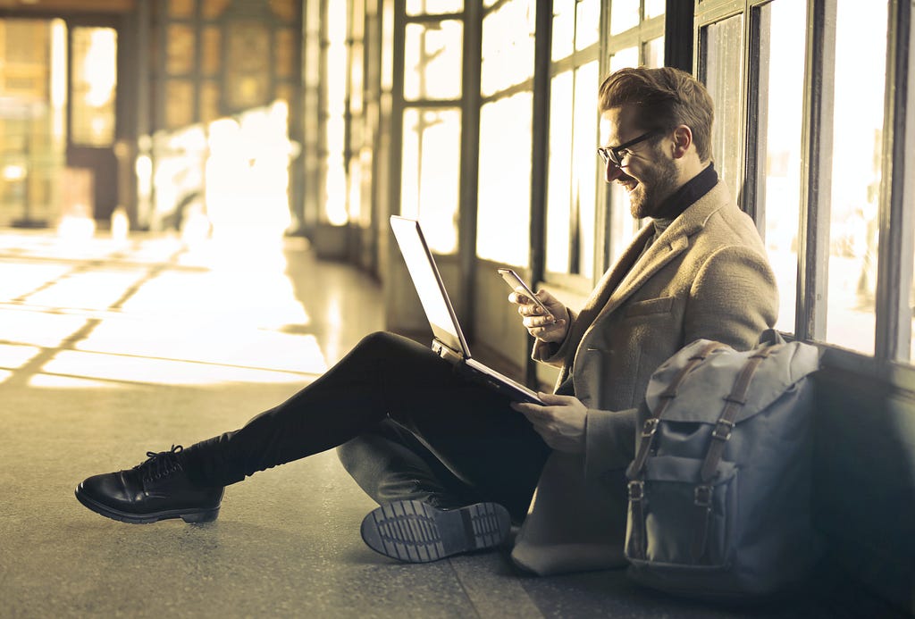 Homem sentado no chão com um notebook no colo e rindo para o celular que está segurando