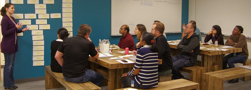 A researcher stands in front of a blue wall covered with sticky notes. An 11-person product team observes the walkthrough.