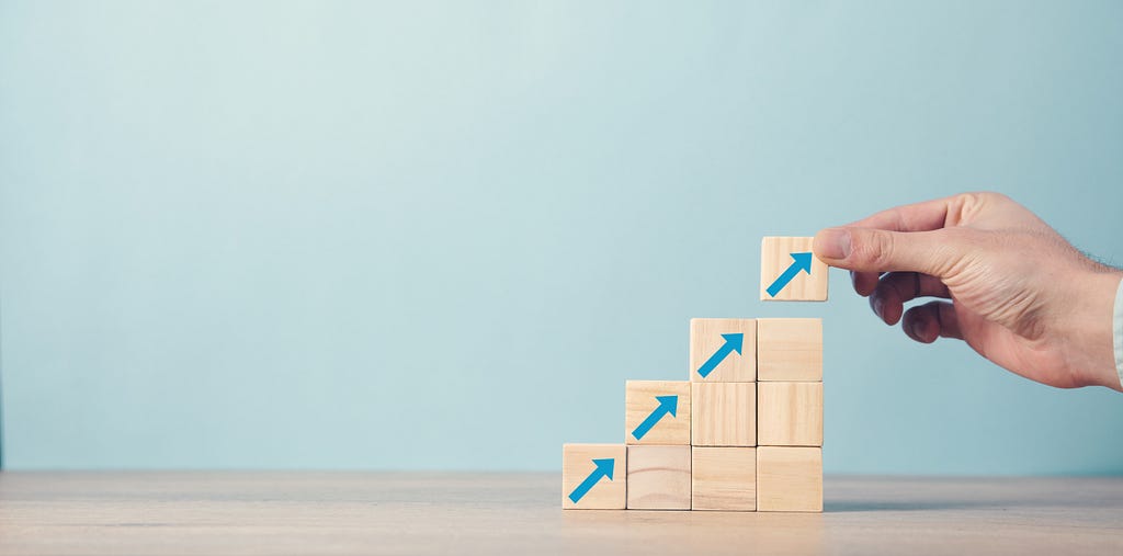 Picture of blocks in a stair-step patter, and on each of the four levels, there’s a block with a blue arrow pointing up and to the right, with the top block with an arrow being placed on the final level by a person’s hand.