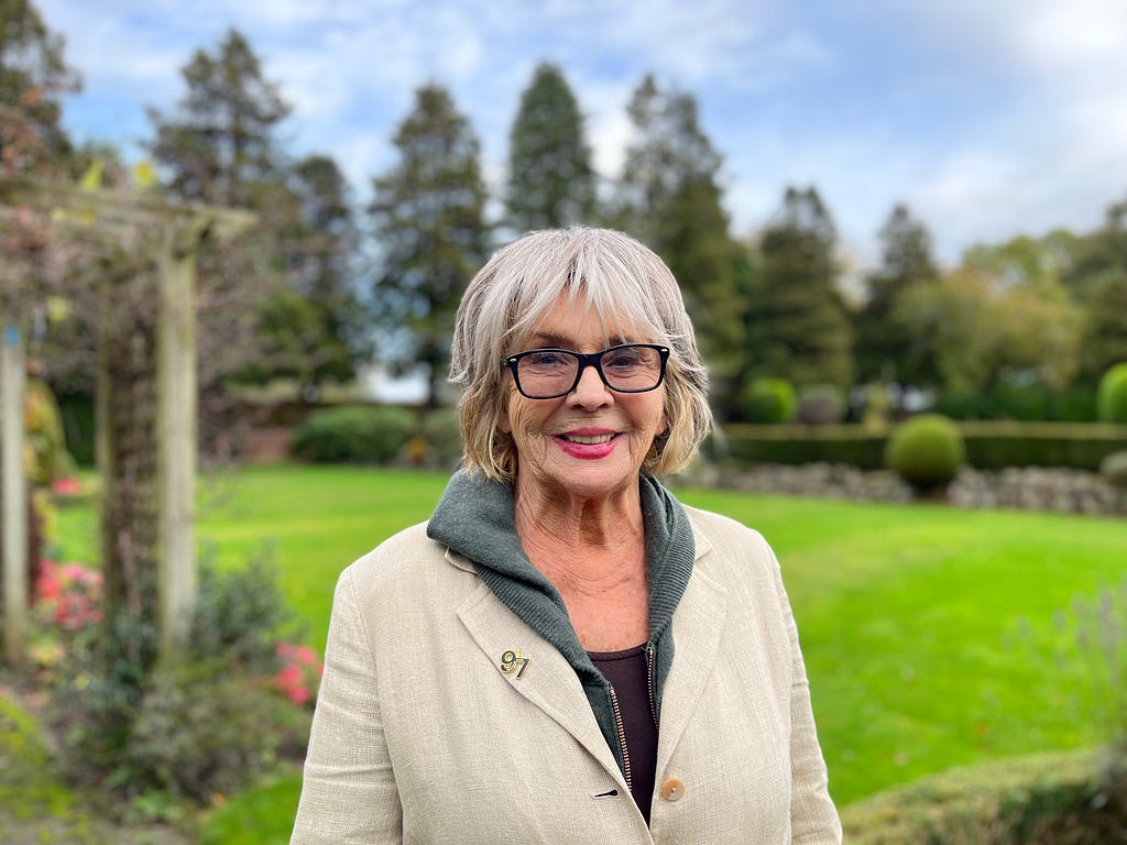 Photograph of actor Sue Johnston stood in her garden. She’s an older woman with short, light-coloured hair and glasses. She’s smiling at the camera.