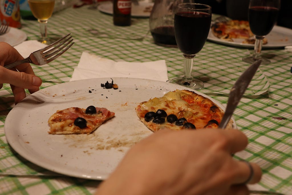 A partially eaten pizza topped with olives, surrounded by a glass of red wine and a simple green-and-white checkered tablecloth, capturing a warm, intimate meal setting.