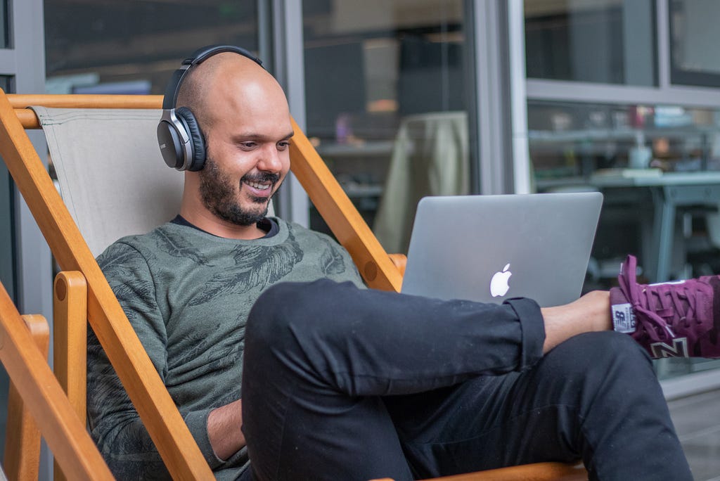 Man sitting on a chair smiling while working on a laptop and using headphones